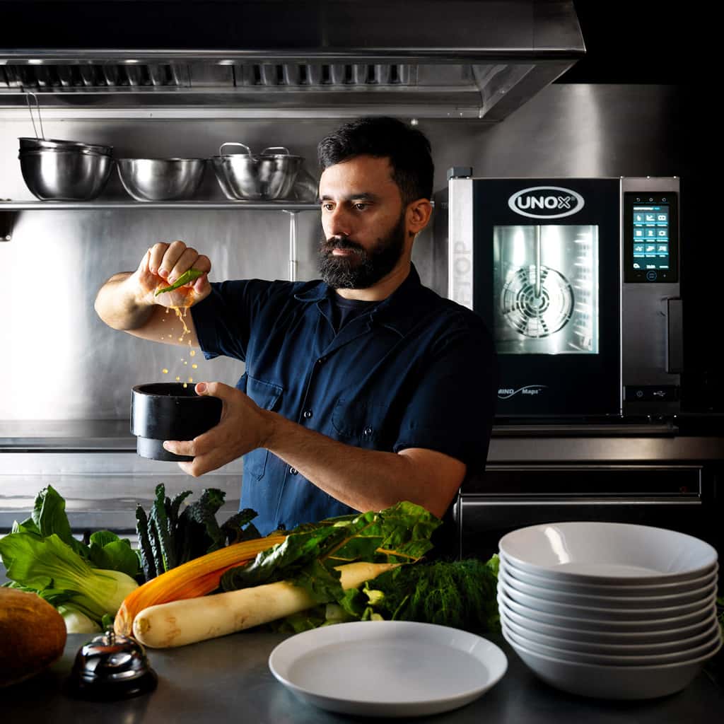 Male chef in kitchen squeezing lime with juice coming out, metal bowls on shelf behind with Unox cheftop combi oven.