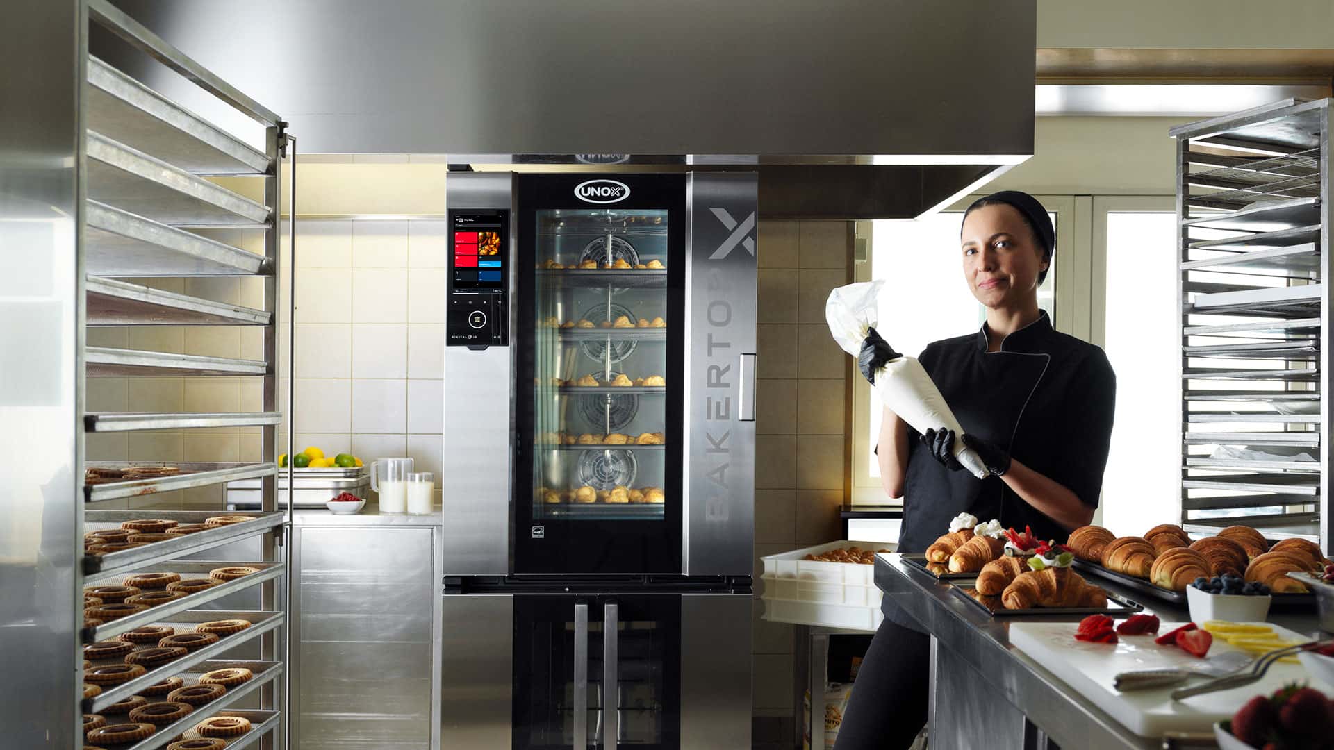 Una pastelera con una manga pastelera delante de una encimera con croissants y un horno mixto de Unox con bollería en su interior a sus espaldas.