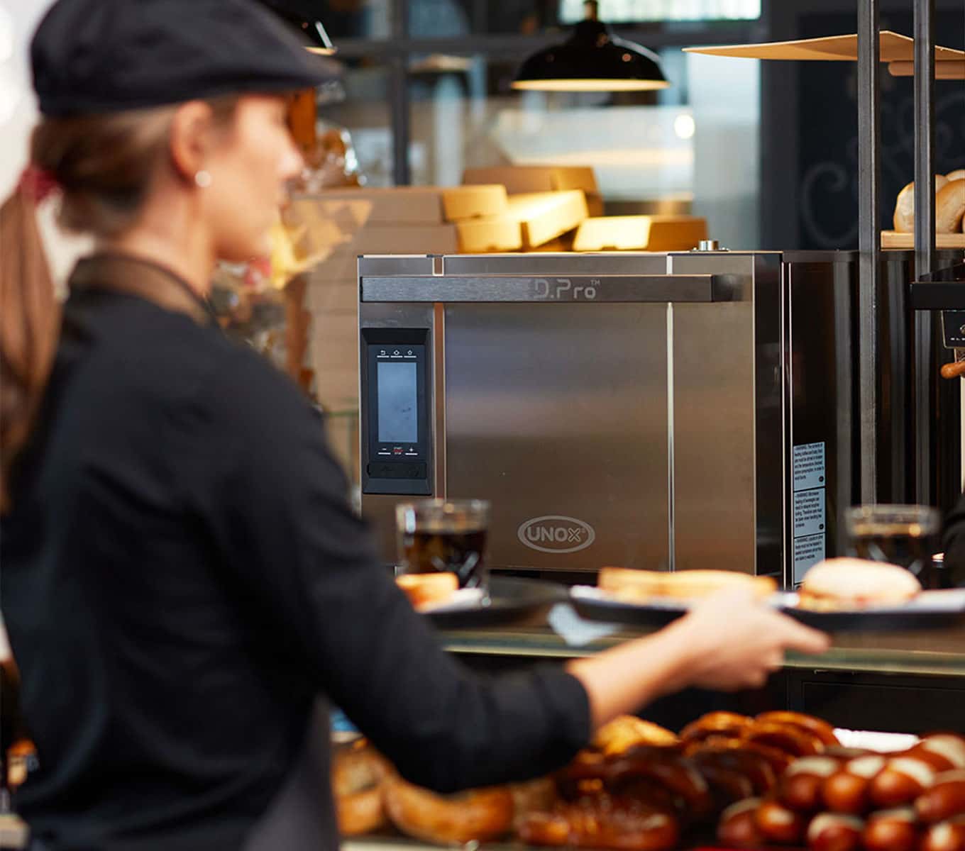 Un’operatrice di bar afferra un vassoio dal banco con un forno rapido professionale Unox SPEED.Pro™ sullo sfondo