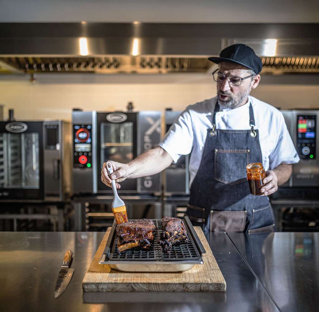 Male chef glazing ribs on a grill, prepared using a Unox combi oven with a professional smoker