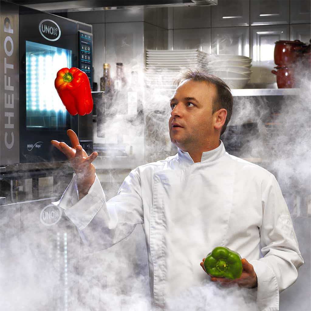 Male chef in white jacket stood in kitchen surrounded by smoke. He is throwing a red pepper in the air, stood next to an Unox combi oven.