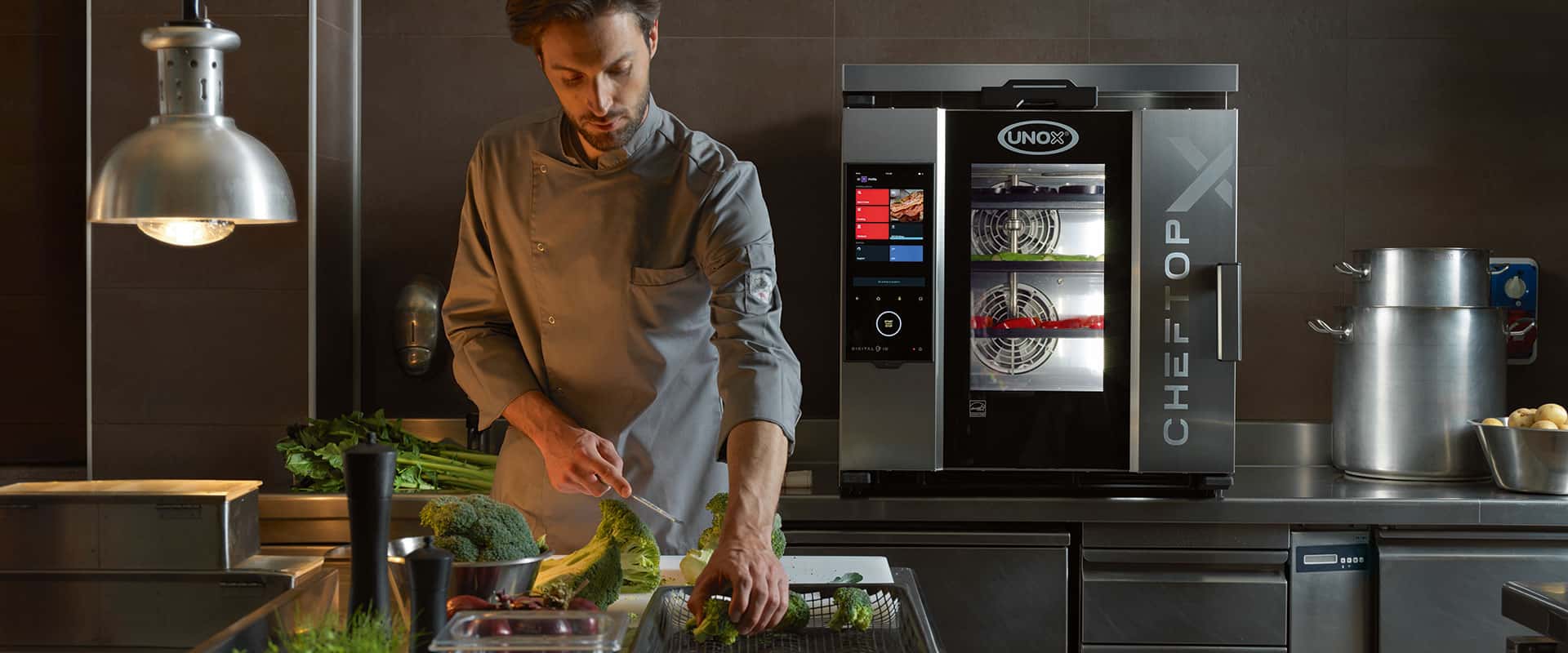 Male Chef standing in a kitchen holding a knife and broccoli, next to an Unox combi oven.