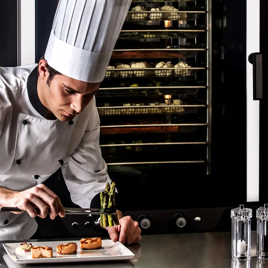 Male chef wearing a chef hat, placing asparagus on plate using tongs, pieces of salmon on plate, Unox oven in background with trays of vegetables.