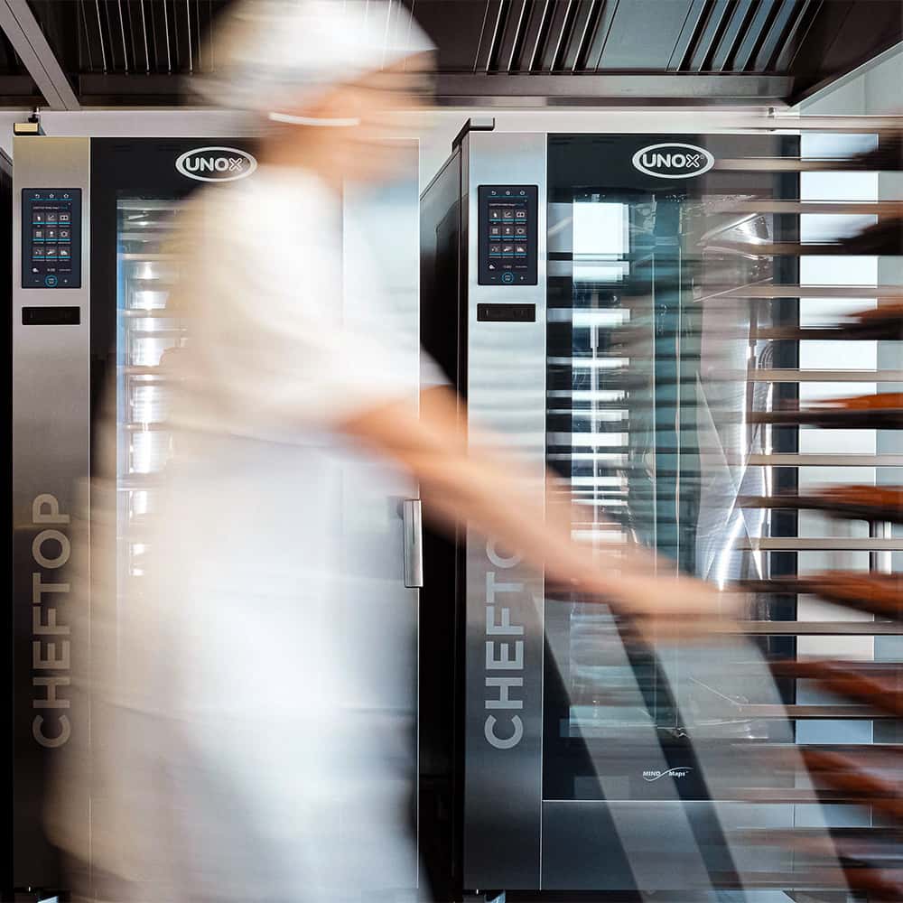 Kitchen with two tall combi trolley ovens next to each other, a chef with a white hat walks past the camera pushing a trolley, causing motion blur.