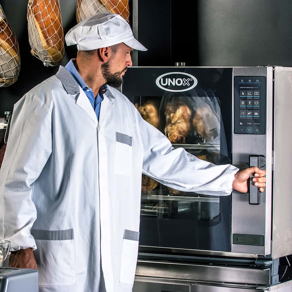 Man in a deli or butchers wearing a white hat, hams hang in the background, opening the door of an Unox combi oven filled with roasted chickens.