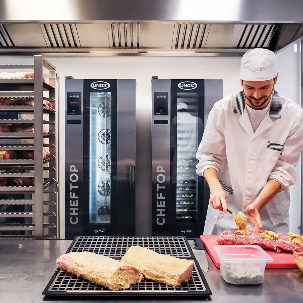 Male chef in kitchen, marinating meat on counter, a tray with two large cuts of meat on counter, two tall combi trolley ovens in the background.