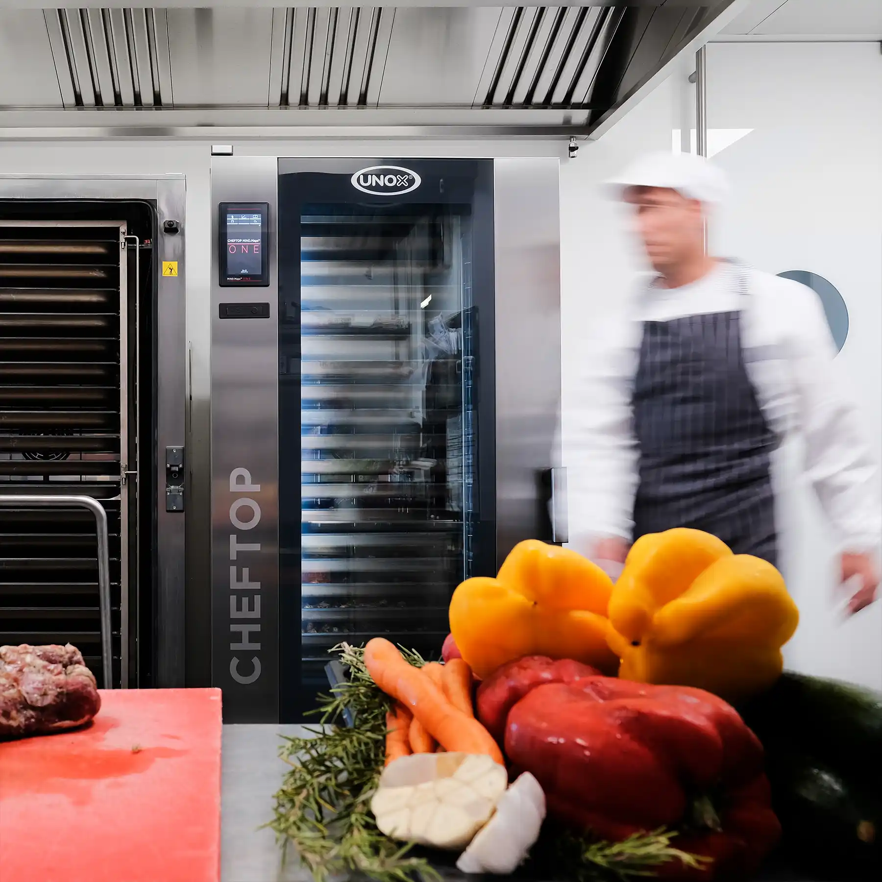 Two tall combi trolley ovens in a kitchen, meat on a red chopping board on counter, alongside vibrant vegetables, male chef walking past oven.