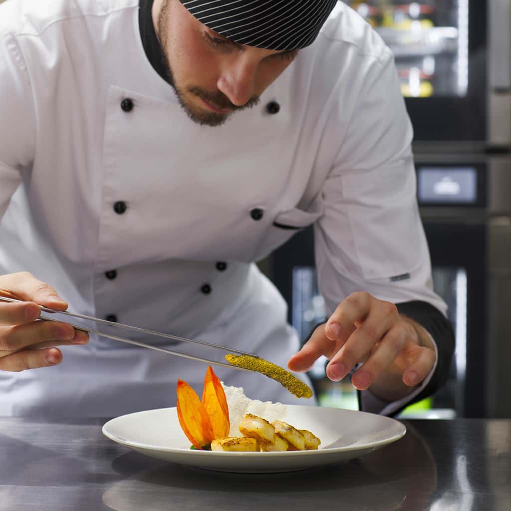 Male chef in kitchen using tongs to plate vegetables on a white plate, Unox Evereo in the background.