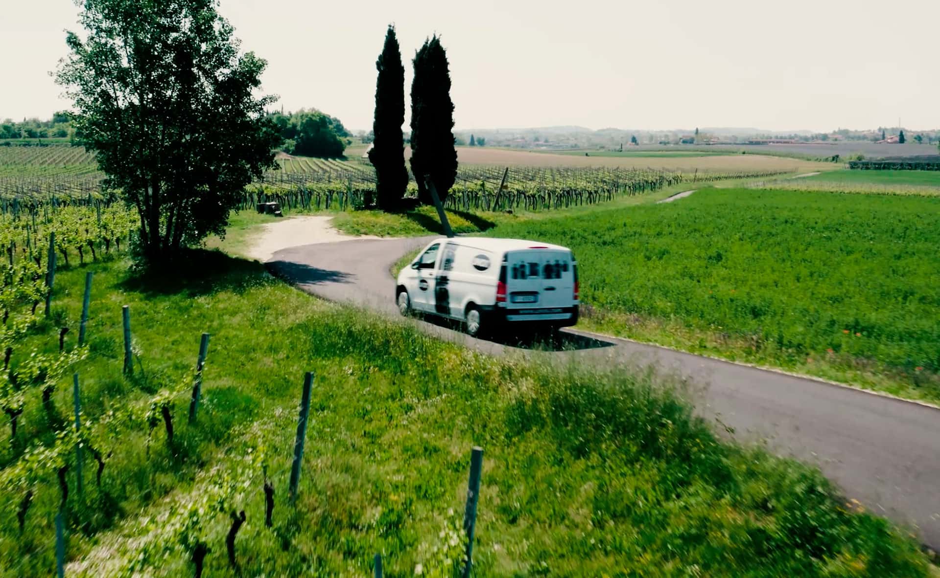 Una furgoneta blanca con el logotipo de Unox e imágenes de hornos impresas circula por una carretera rodeada de campos verdes y árboles.
