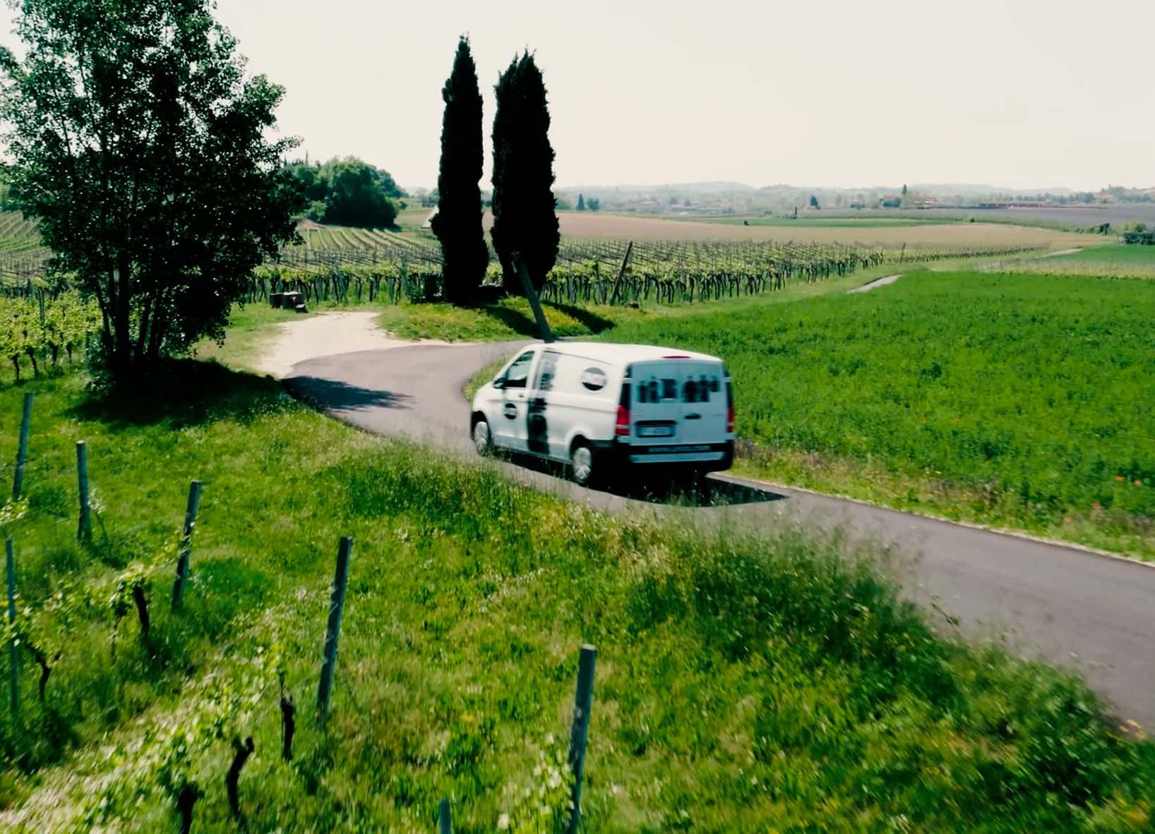 Una furgoneta blanca con el logotipo de Unox e imágenes de hornos impresas circula por una carretera rodeada de campos verdes y árboles.