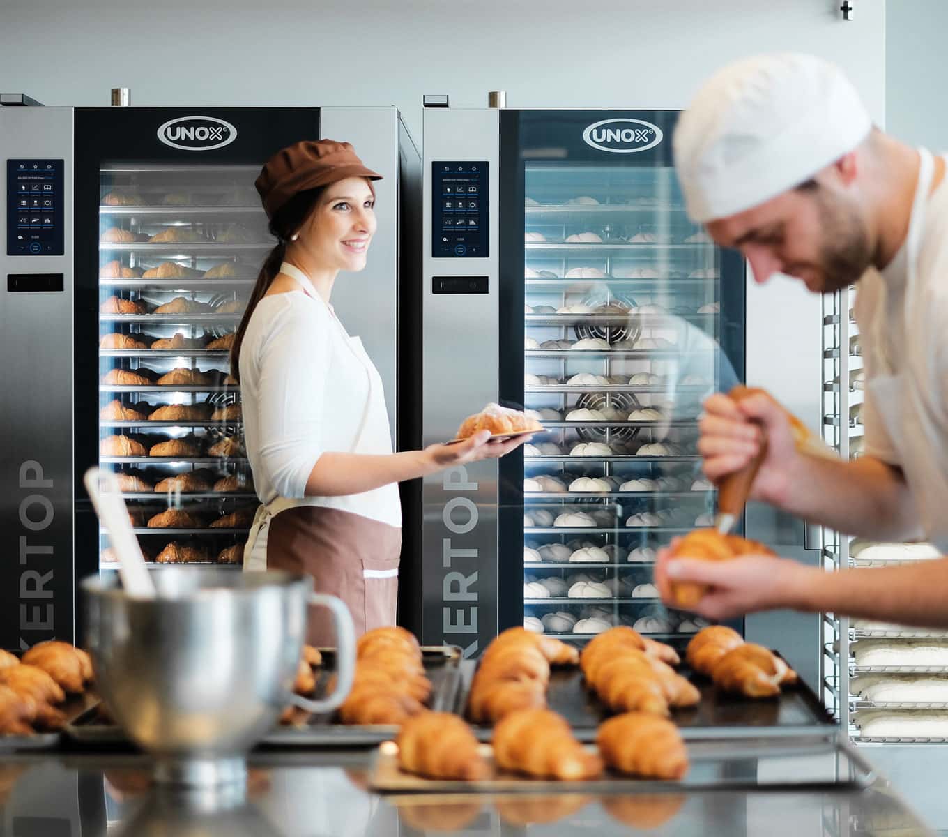 Due operatori preparano croissant in laboratorio con sullo sfondo due forni combinati carrellati Unox a pieno carico