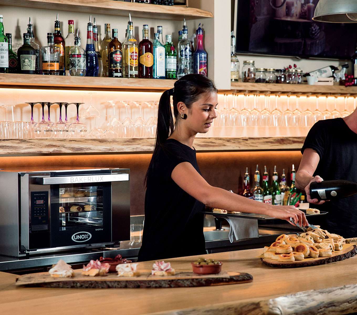 A male operator pours a drink into a glass while a female operator fills a tray with mini pizzas next to a Unox small commercial oven behind the counter