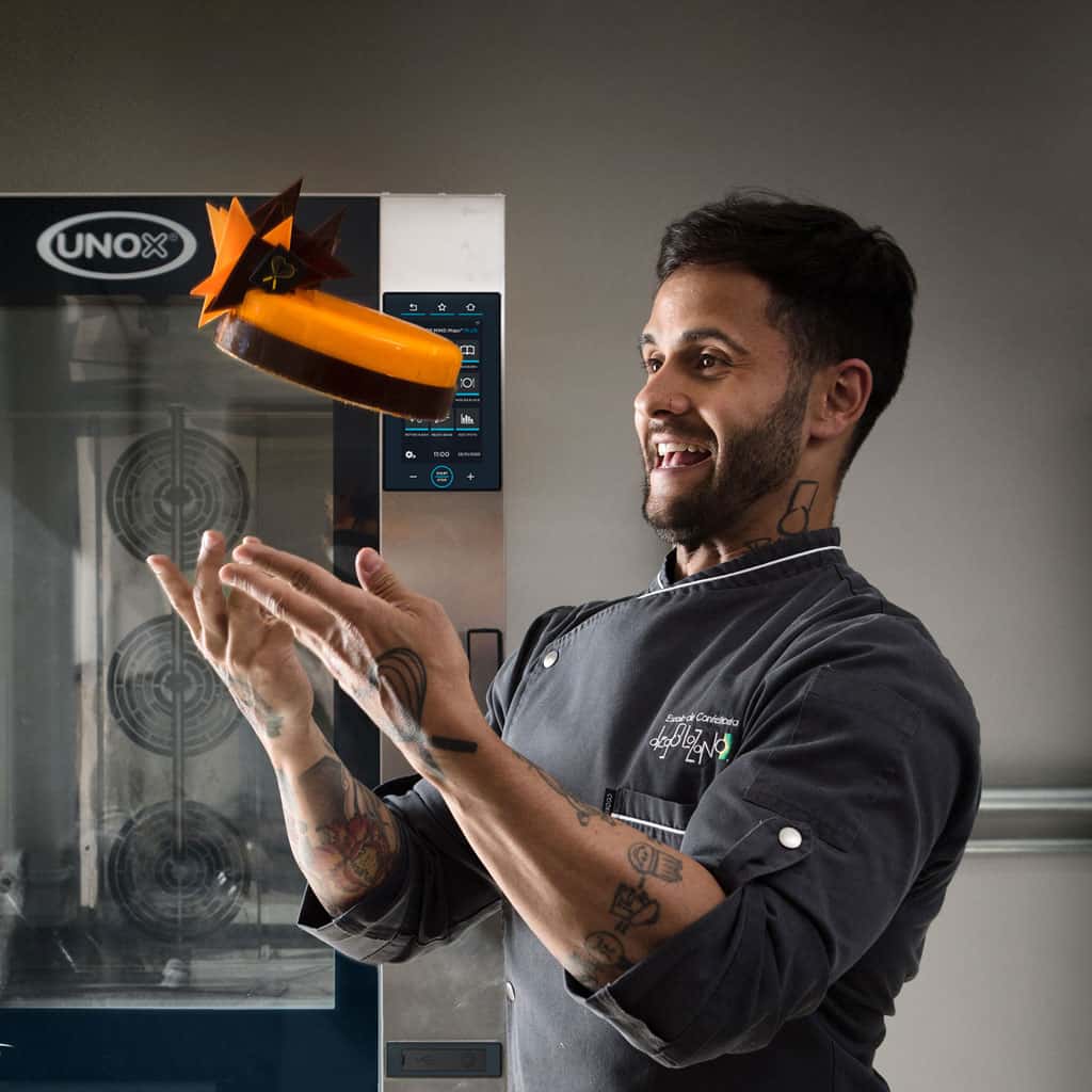 Smiling male chef in kitchen throwing an orange cake into the air, in front of an Unox baking oven.