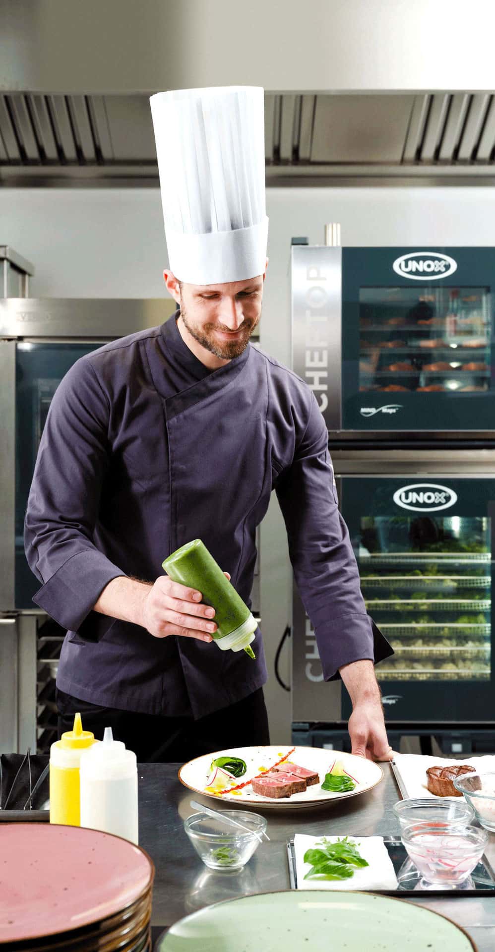 Smiling man in kitchen, wearing chef hat, holding green bottle of sauce over a plate of food, Unox cheftop combi ovens behind him. 