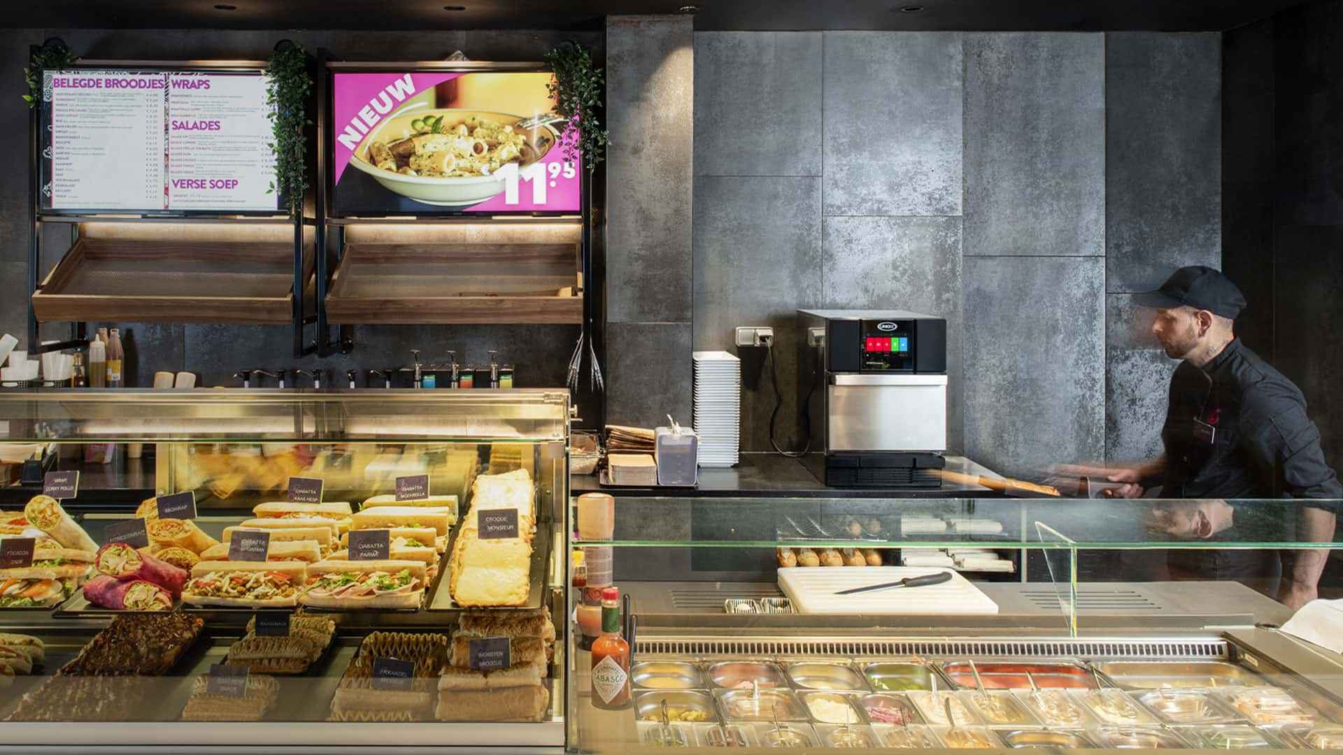 Operador de una estación de servicio mirando un horno rápido Unox detrás de un mostrador con wraps, bocadillos y ensaladas.