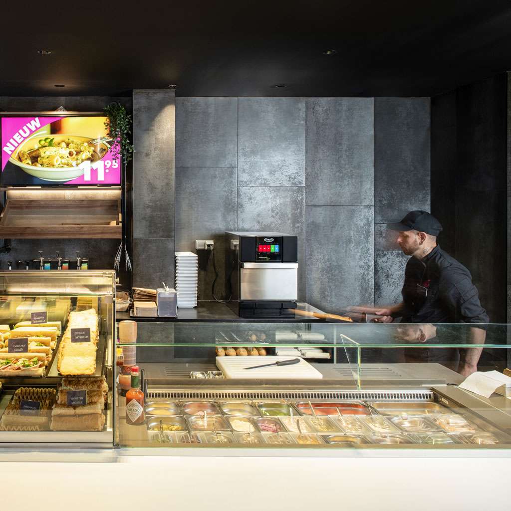 QSR male operator looking at an Unox speed oven behind a counter with wraps, sandwiches, and salads.