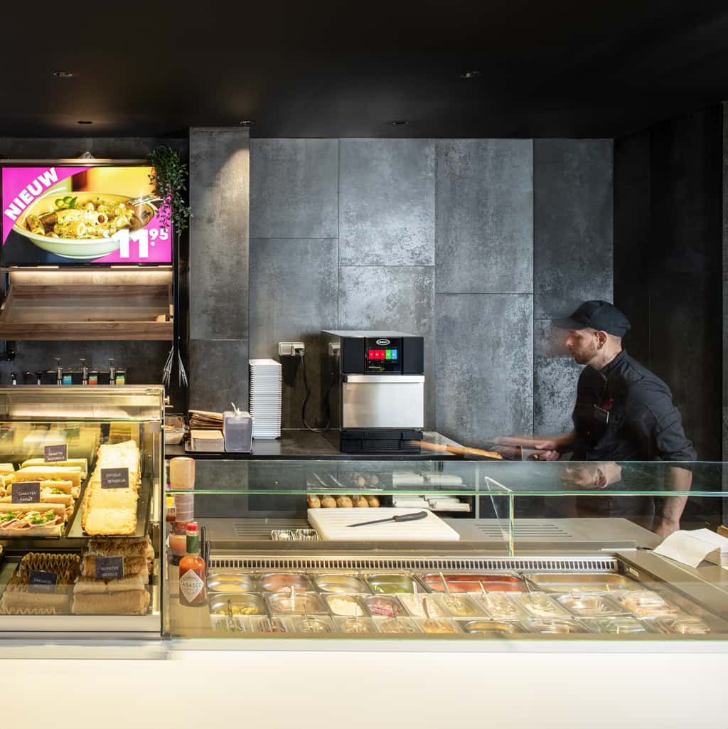 Operador de una estación de servicio mirando un horno rápido Unox detrás de un mostrador con wraps, bocadillos y ensaladas.