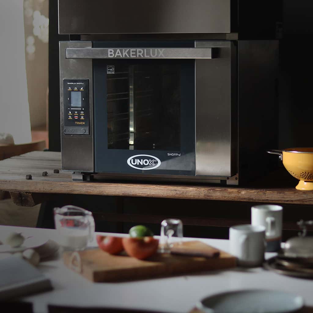 Unox baking oven on a wooden table, countertop on front has sliced tomato on chopping board and various cups and mugs.