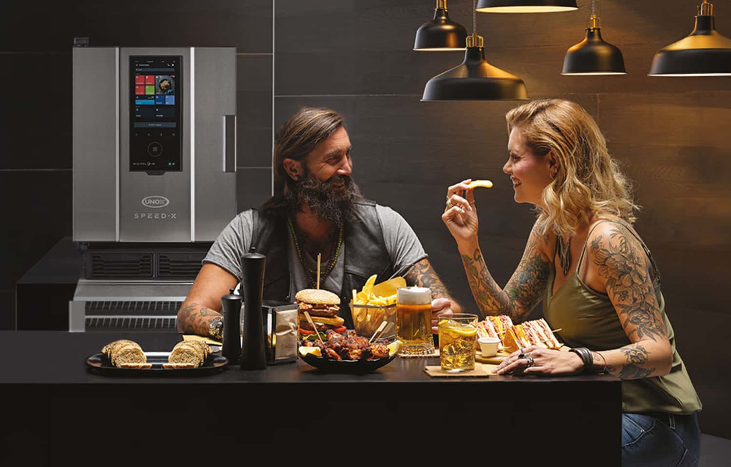 A smiling man and woman with tattoos sit at a table with burgers, chips, sandwich and other food, beer, a cocktail and an Unox speed oven behind them.