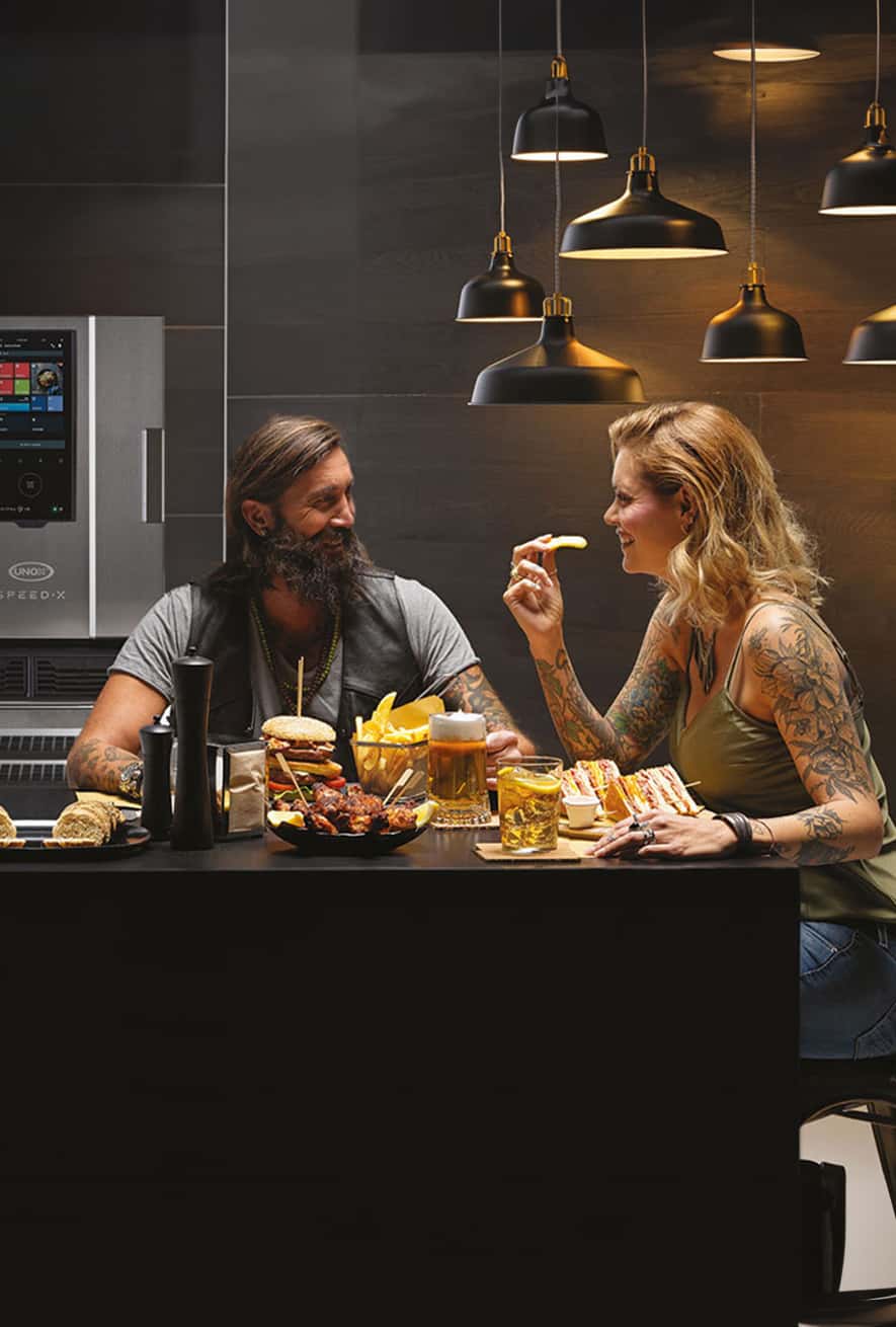 A smiling man and woman with tattoos sit at a table with burgers, chips, sandwich and other food, beer, a cocktail and an Unox speed oven behind them.