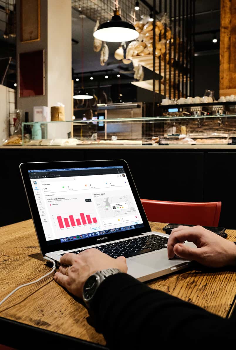 Man sat at table in café working on a laptop, graphs showing data can be seen on screen.