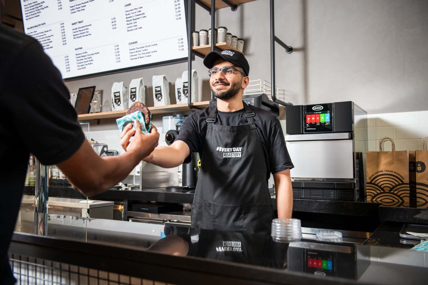 Un barista sirve una galleta caliente a un cliente, con un horno SPEED.Compact de Unox visible sobre la encimera.