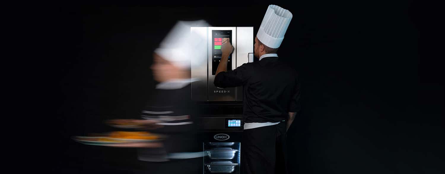 Male chef touching display panel on speed oven, stacked on top of an Unox Evereo, a chef walking fast holding plates of food.