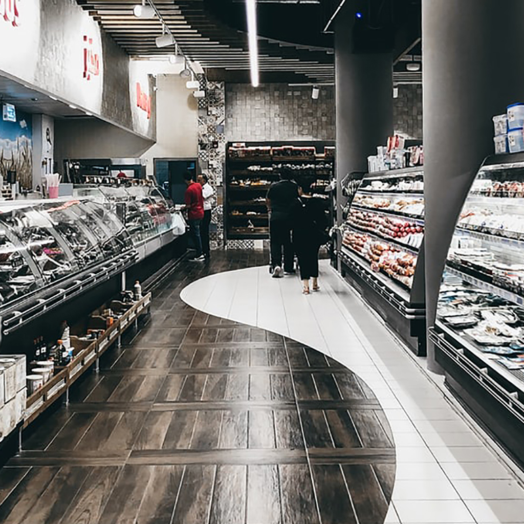 Supermarket aisle with deli counter on one side and chilled cabinet on the other side. Customers can be seen pushing a trolley.